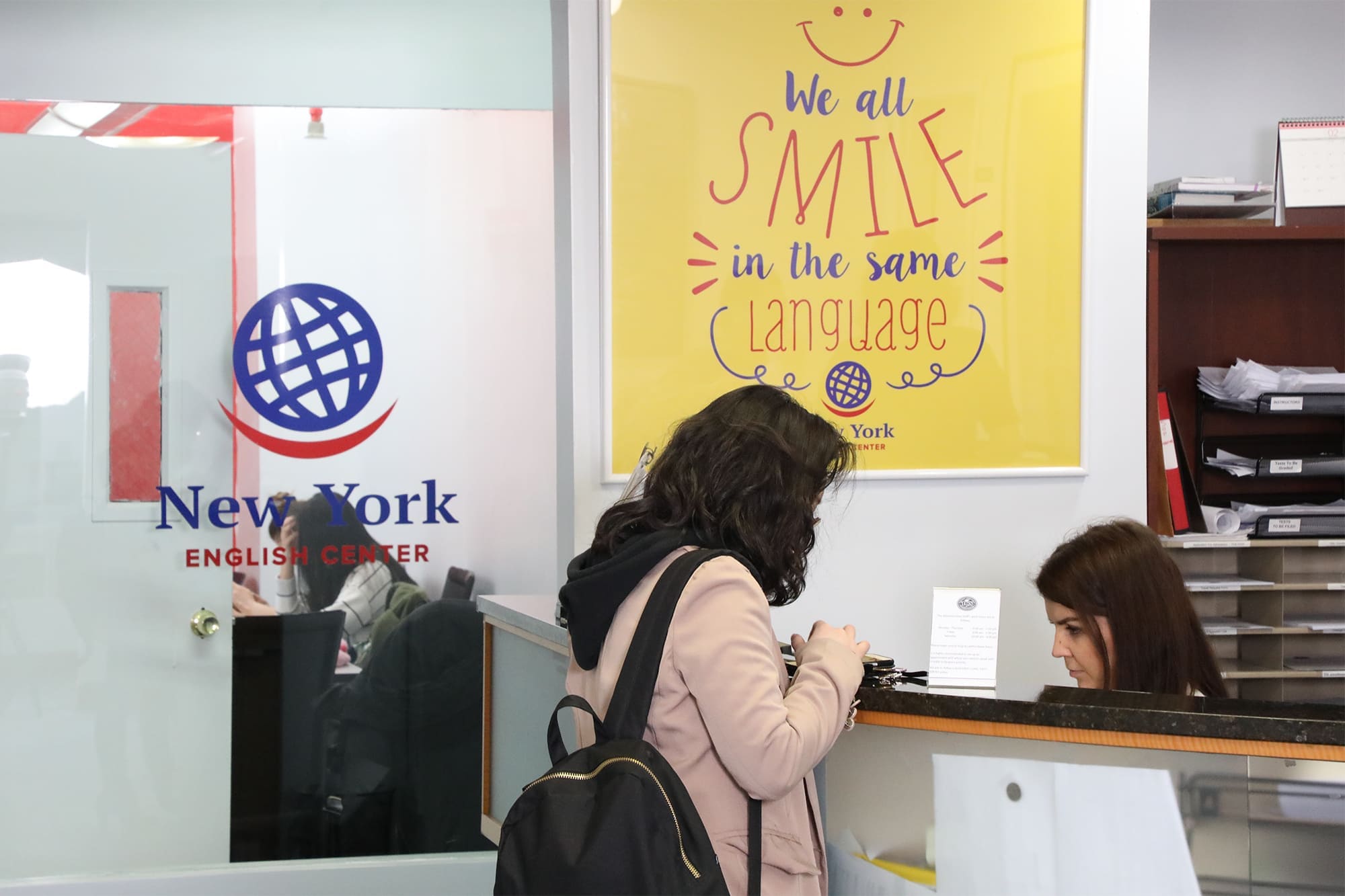 Two women at the front desk of New York English Center's reception area, engaged in conversation.